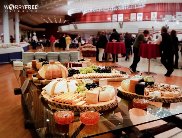 A long glass table filled with catering food including cheese platters, grapes, and appetizers in a large event hall.