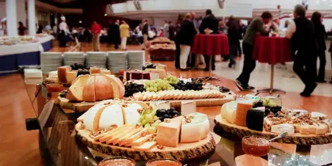 A long glass table filled with catering food including cheese platters, grapes, and appetizers in a large event hall.