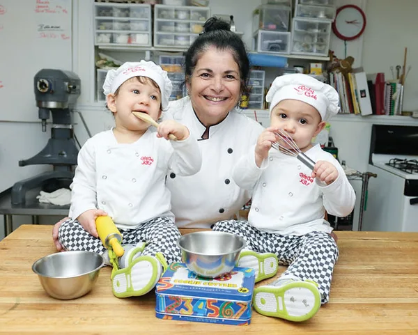 A woman in a chef's coat sitting at a kitchen table with two young children wearing chef hats, surrounded by baking ingredients and equipment.