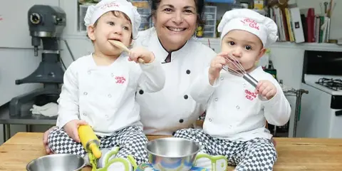 A woman in a chef's coat sitting at a kitchen table with two young children wearing chef hats, surrounded by baking ingredients and equipment.