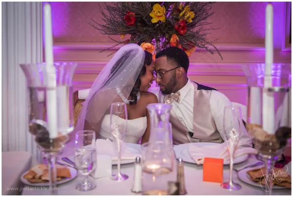Wedding couple seated at a reception table with purple uplighting on the wall behind them and floral centerpiece.