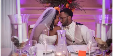 Wedding couple seated at a reception table with purple uplighting on the wall behind them and floral centerpiece.