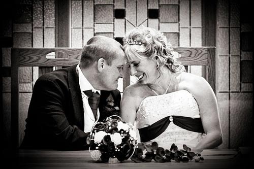 A black and white photograph of a bride and groom smiling at each other during a wedding ceremony.