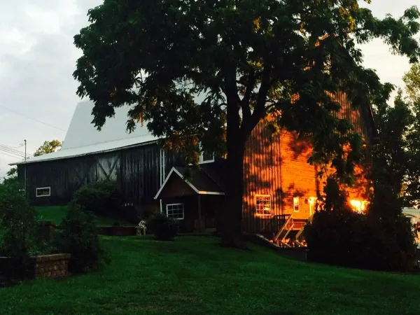 Exterior view of a large historic barn with a gambrel roof and attached entryway, surrounded by a grassy lawn and trees at dusk.