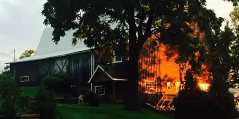 Exterior view of a large historic barn with a gambrel roof and attached entryway, surrounded by a grassy lawn and trees at dusk.