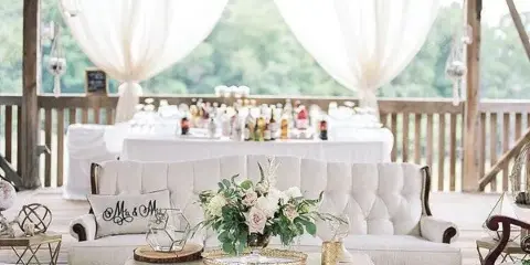 A wedding lounge setup with a white sofa, coffee table, and floral centerpiece in a rustic venue.