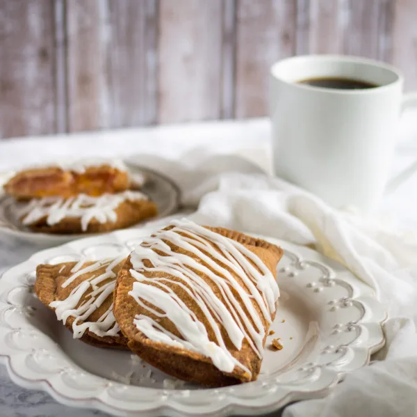 Two golden-brown fried pies drizzled with white icing on a white plate, with a cup of coffee in the background.