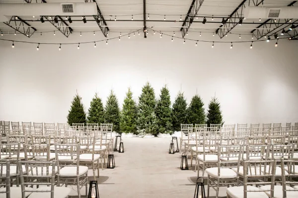A wedding ceremony setup with rows of white chairs facing a backdrop of evergreen trees and string lights.