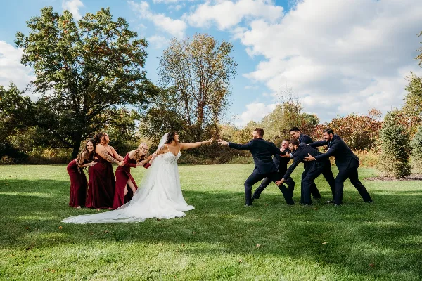 Bride in a white gown and groomsmen in dark suits playfully reach toward each other on a grassy lawn with bridesmaids in red dresses and trees in the background.