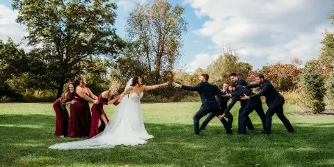 Bride in a white gown and groomsmen in dark suits playfully reach toward each other on a grassy lawn with bridesmaids in red dresses and trees in the background.
