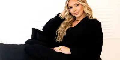 Portrait of a blonde woman in a black suit sitting on a chair, showcasing her makeup and hair.