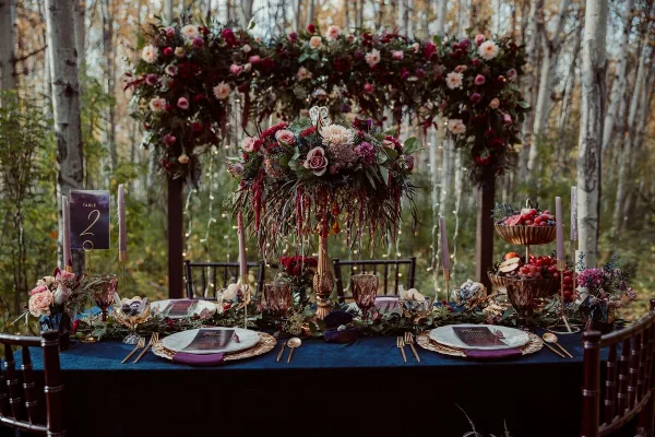 A wedding reception table set with a floral arch, candles, and place settings in a forest venue.