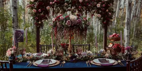 A wedding reception table set with a floral arch, candles, and place settings in a forest venue.