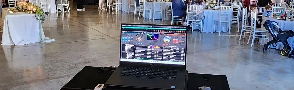 A laptop running DJ software set up in a wedding reception hall with tables and chairs in the background.