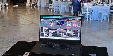 A laptop running DJ software set up in a wedding reception hall with tables and chairs in the background.