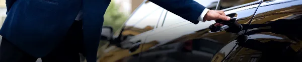 A professional chauffeur in a dark suit opening the door of a black luxury sedan.