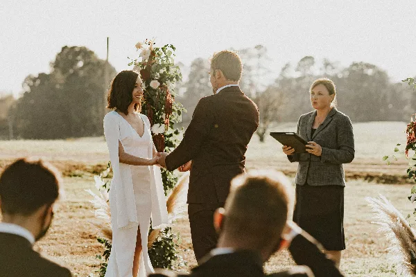 A female officiant in a grey blazer stands holding a tablet while officiating an outdoor wedding ceremony for a bride and groom.