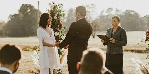 A female officiant in a grey blazer stands holding a tablet while officiating an outdoor wedding ceremony for a bride and groom.