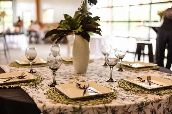 A set dining table with floral centerpiece and glassware inside a bright event venue with large glass doors.